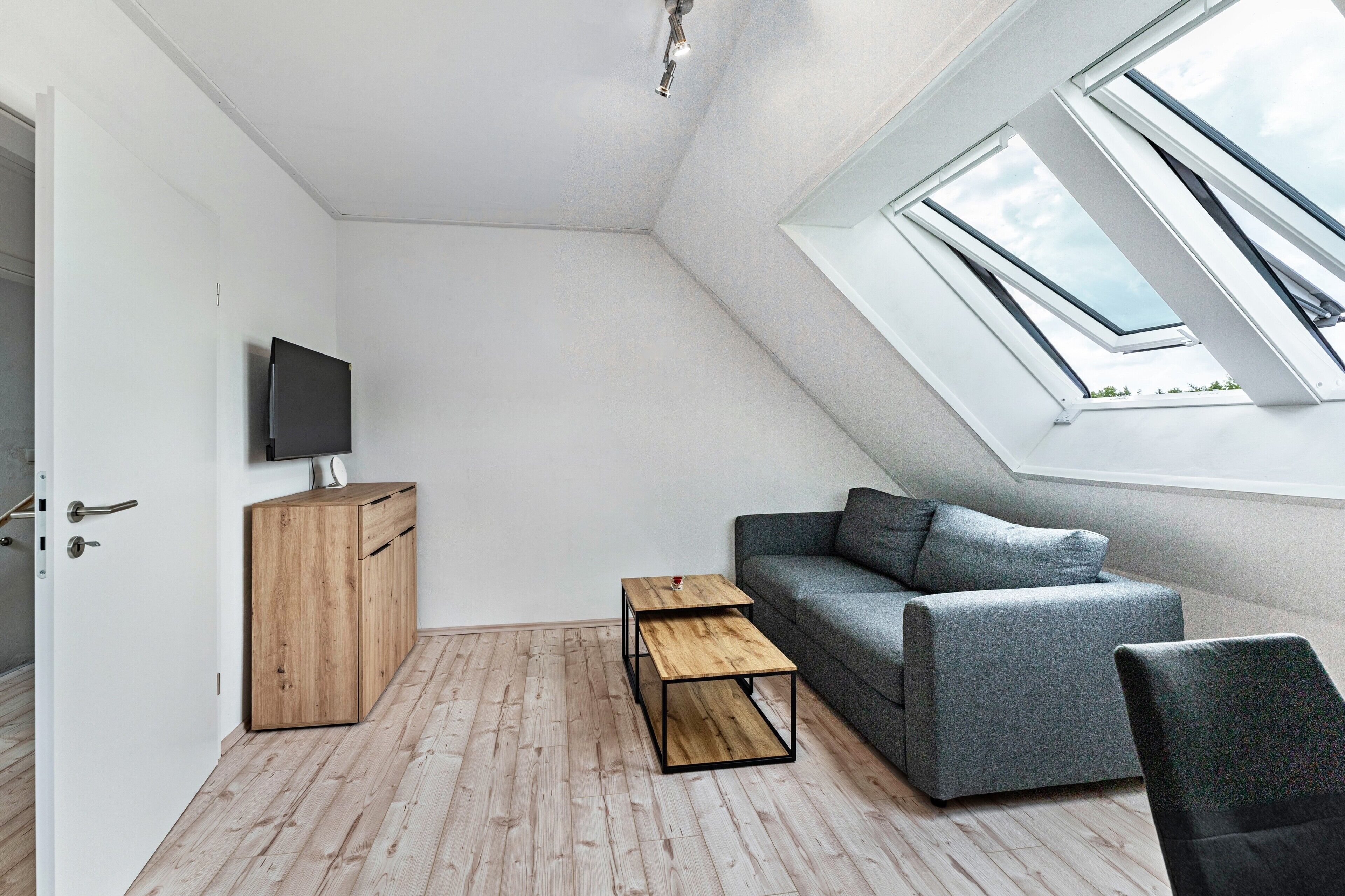 A room with a sloped ceiling, a wooden cabinet, a gray sofa, a small wooden table, and a TV mounted on the wall.