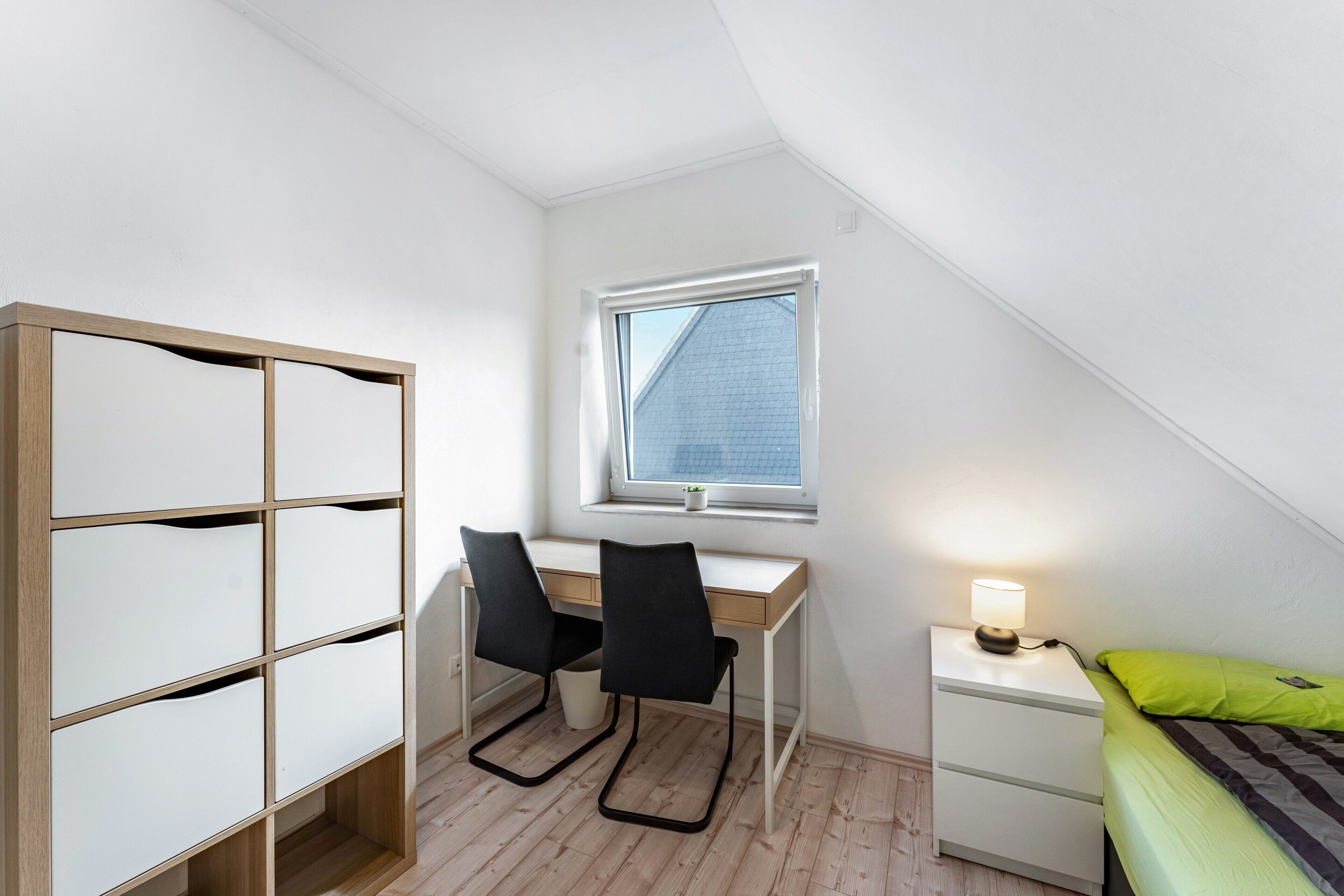 Ein Schlafzimmer mit Schreibtisch, zwei Stühlen, einem Nachttisch mit Lampe, einem Bett mit grünem Kissen und einem Fenster mit Blick auf ein Gebäude.
