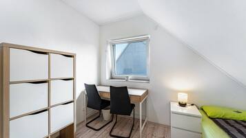 Ein Schlafzimmer mit Schreibtisch, zwei Stühlen, einem Nachttisch mit Lampe, einem Bett mit grünem Kissen und einem Fenster mit Blick auf ein Gebäude.