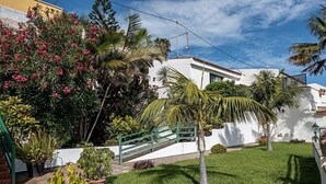 A well-maintained garden with a variety of plants and a palm tree, a white building with a red-tiled roof, and a green metal staircase leading up to the building. - Apartments with terraca inside of banana plantation with ocean view (Garachico)