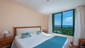 A double bed with a blue blanket, two nightstands with lamps, a window with a view of the ocean, and a balcony with a green railing. - Apartments with terraca inside of banana plantation with ocean view (Garachico)