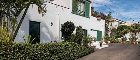 A white building with green shutters, a paved driveway, and lush greenery.