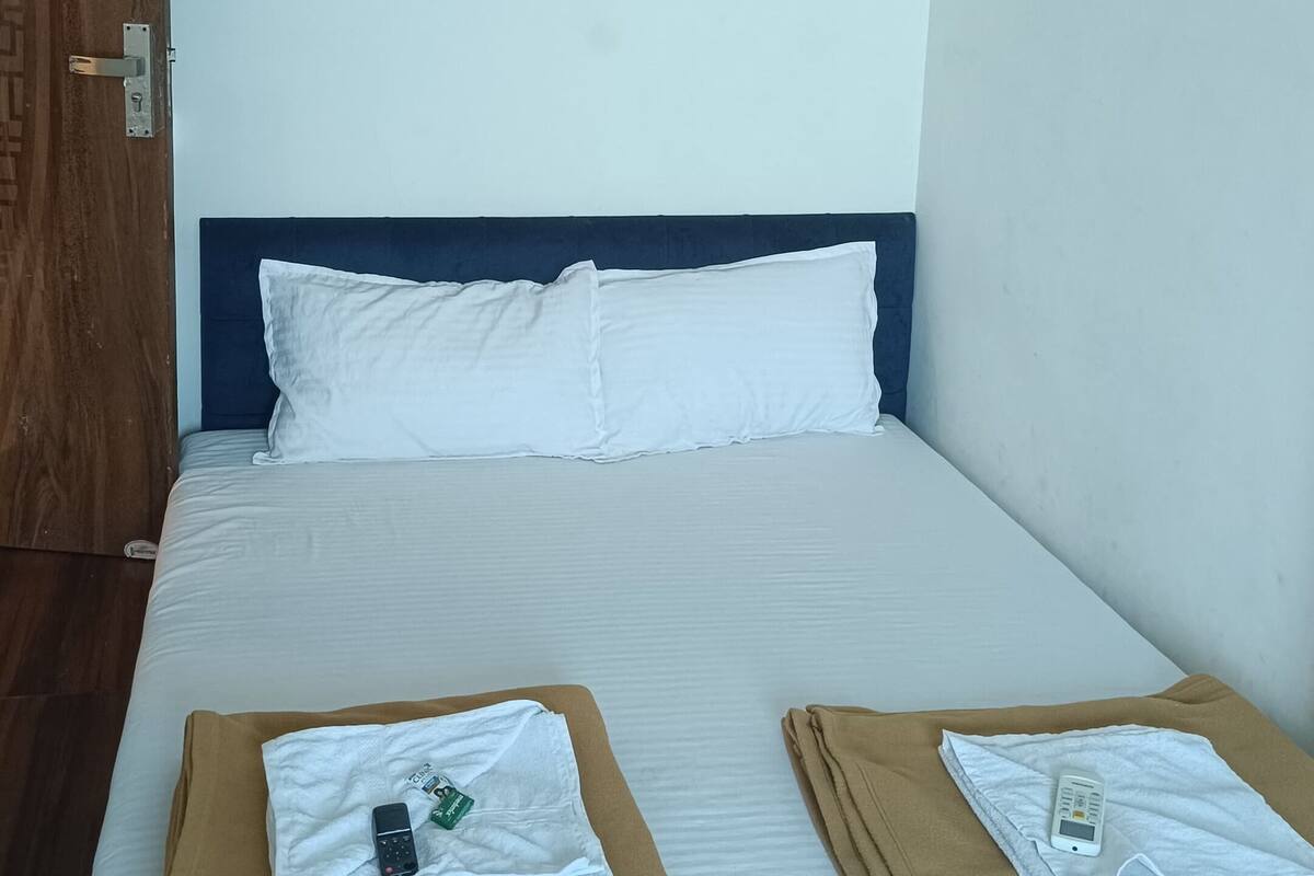 A neatly made bed with white linens, a dark blue headboard, and two folded brown towels with remote controls on them.