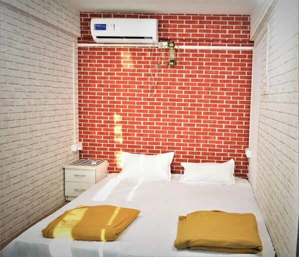 A room with a brick-patterned wall, a bed with white bedding, and a white air conditioning unit above it.