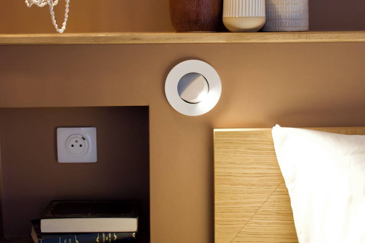 Una habitación ordenada con cabecera de madera, una mesita de noche con lámpara, y una estantería con libros y objetos decorativos.