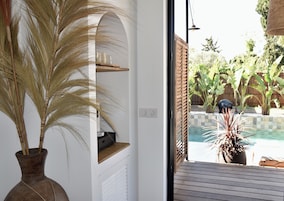 Una habitación con una planta en maceta grande, un estante con cafetera y vistas a una piscina y jardín exterior.