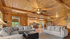 A spacious living room with a large sectional sofa, a matching armchair, and a coffee table. A wooden ceiling fan and a staircase with a wooden railing are visible. - Private 2+ Acre Ridge-Top Retreat with Hot Tub, Game Room & Stunning Views! (Gatlinburg)