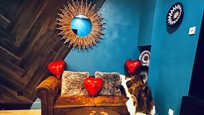 A cozy room with a wooden herringbone floor, a blue wall, a round mirror, and a brown sofa with heart-shaped pillows.