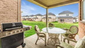 Een terras buiten met een barbecue, rieten stoelen en een glazen tafel.
