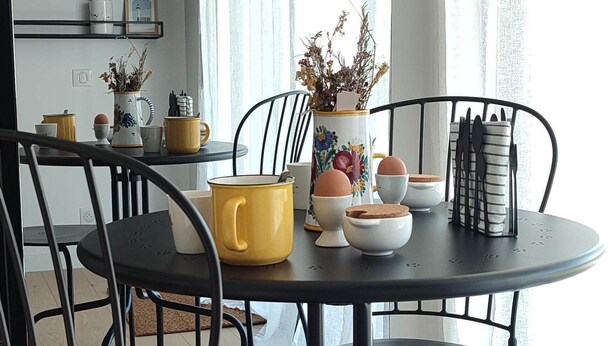 Un coin repas avec une table ronde, des chaises noires et une tasse jaune. On aperçoit en arrière-plan une cuisine avec un meuble noir et une étagère ornée d’objets décoratifs.