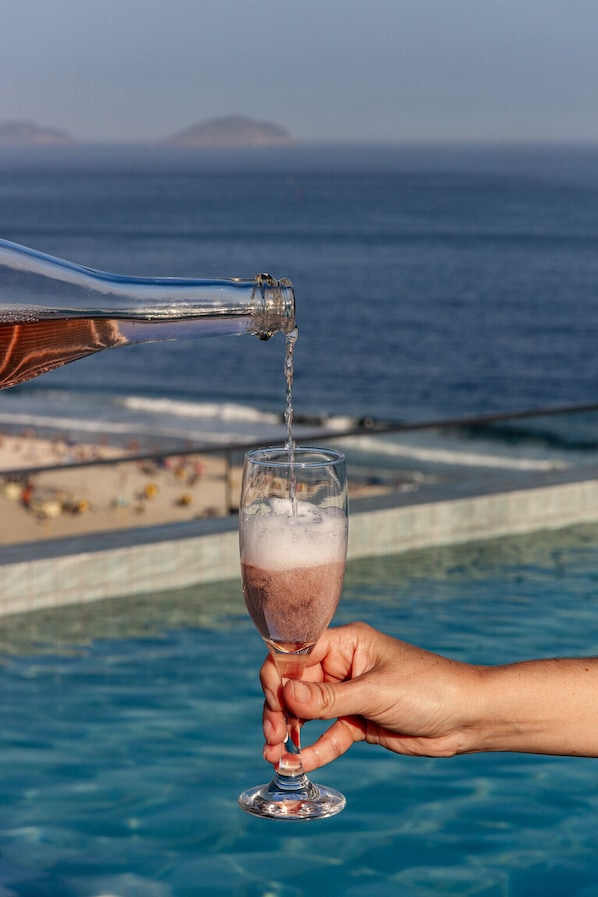 A hand pouring a drink into a glass by a pool with a beach and ocean in the background.