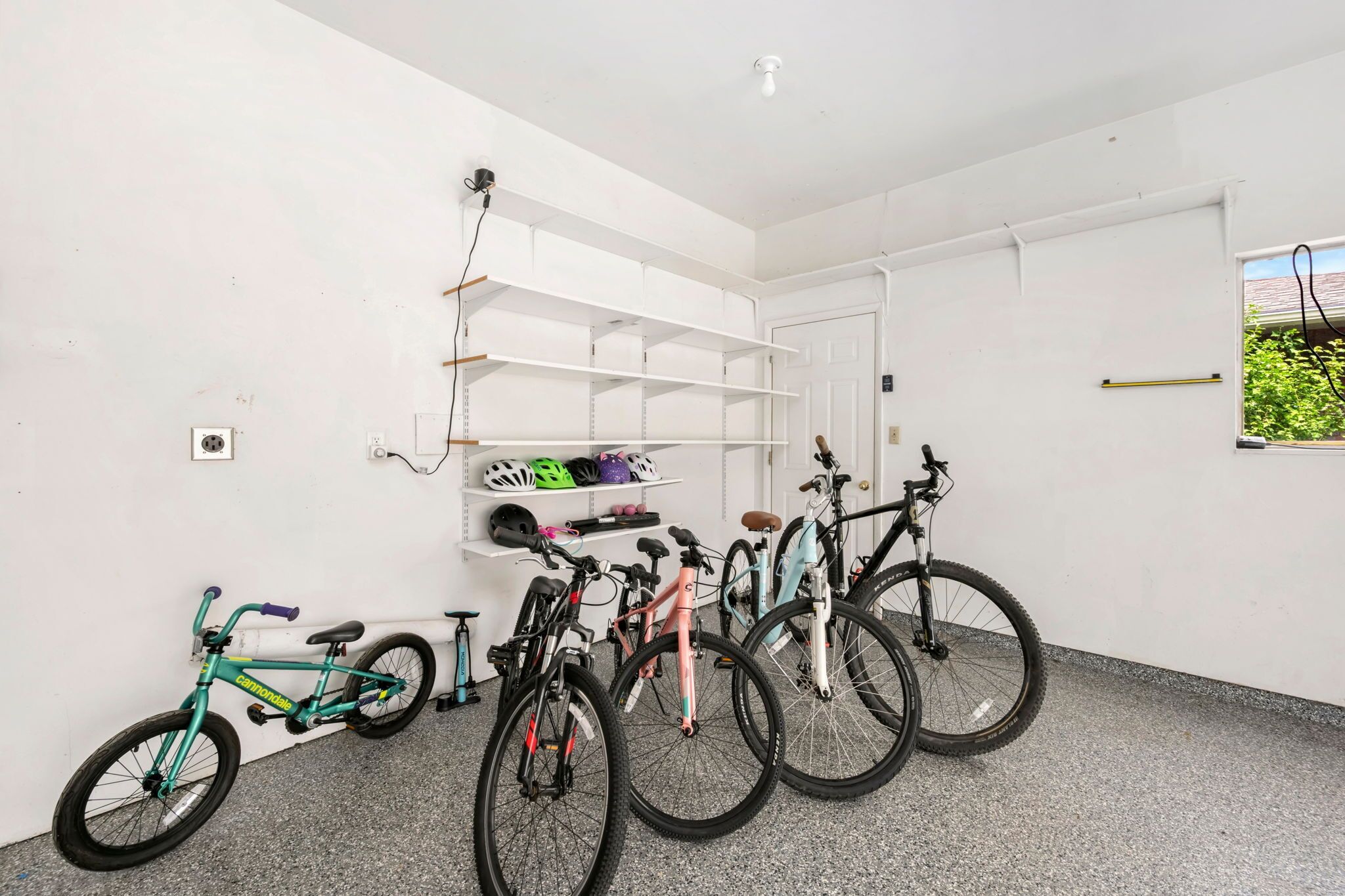 Une pièce aux murs blancs, avec une porte et une fenêtre. Il y a des vélos et des casques sur le sol. Des étagères sont fixées au mur.