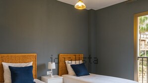 Ein Hotelzimmer mit zwei Betten, jeweils mit weißer Bettdecke und blauen Kissen, einem Rattan-Kopfstück, einem Nachttisch mit Lampe und einem Fenster mit Blick ins Grüne.
