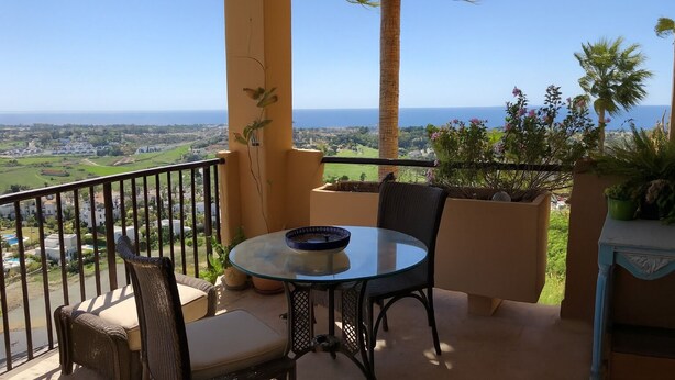 Ein Balkon mit einem runden Glastisch und zwei Stühlen, mit Blick auf eine Küstenlandschaft mit Palmen und Meer.