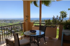 A balcony with a round glass table and two chairs, overlooking a coastal landscape with palm trees and a view of the sea.
