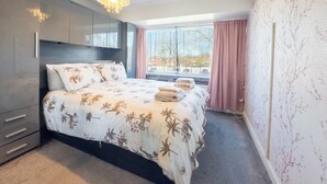 Une chambre à coucher avec un lit, une armoire, un lustre, une fenêtre avec des rideaux et une vue sur les arbres.