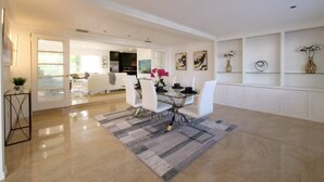 Un comedor amplio con una mesa de vidrio y sillas blancas, una cocina al fondo y un pasillo que lleva a una sala de estar.