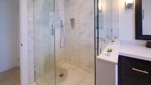 Un baño moderno con ducha envuelta en vidrio, piso de mármol blanco y tocador blanco con gabinete negro.