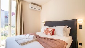 A hotel room with a bed, a window with curtains, an air conditioning unit, and a wall-mounted light fixture.