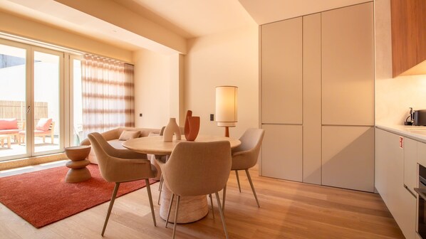 A modern living room with a round table, comfortable seating, a large window with curtains, and a kitchen area with cabinets.