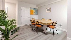 Uma sala de jantar com uma mesa retangular e seis cadeiras, um vaso com planta, uma porta branca e um quadro na parede.