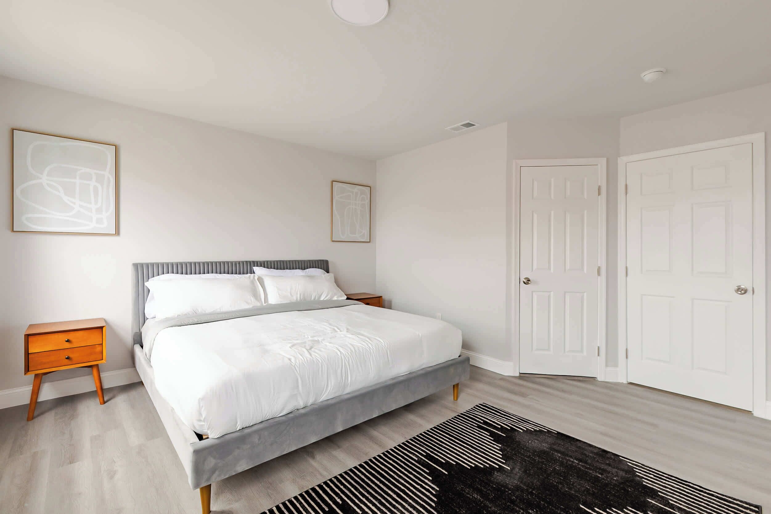 Een moderne slaapkamer met een grijs bed, witte beddengoed, een houten nachtkastje, een zwart-wit tapijt en twee abstracte schilderijen in lijsten aan de muur.
