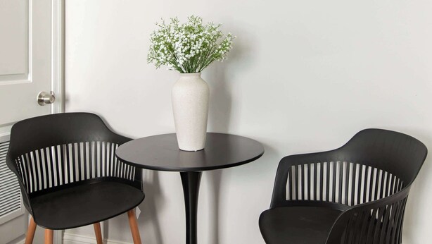 Ein Zimmer mit einem runden schwarzen Tisch, zwei schwarzen Stühlen mit Holzbeinen, einer weißen Vase mit Blumen und einem farbenfrohen abstrakten Gemälde an einem hölzernen Regal über dem Tisch.