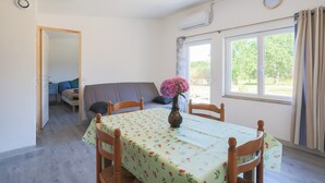 A dining area with a table, chairs, a vase with flowers, a clock, and a window with curtains. - Holiday Home 'Glycine' in Paimpol with Private Kitchen, Air Conditioning, and Bike Storage (Paimpol)