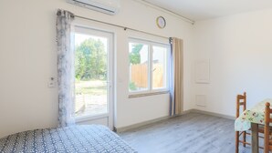A room with a bed, a table with a floral tablecloth, a window with curtains, and an air conditioning unit. - Holiday Home 'Glycine' in Paimpol with Private Kitchen, Air Conditioning, and Bike Storage (Paimpol)