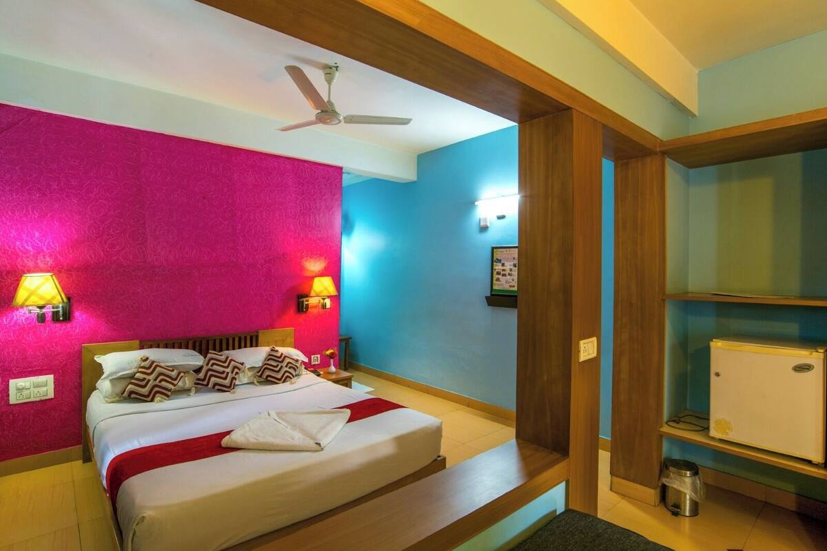 A bedroom with a bed, two bedside lamps, a ceiling fan, a small refrigerator, and a wall-mounted shelf.