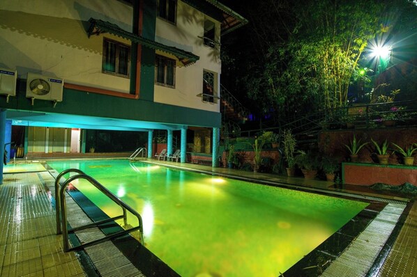 A swimming pool with a tiled deck, a metal ladder, and a building with multiple windows and air conditioning units in the background. - Harithagiri Hotel (Kalpetta)