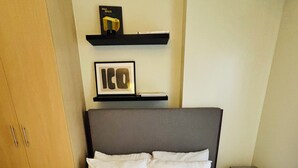 A neatly made bed with white linens in a room with a wooden wardrobe and a shelf with books. - Nature Lovers' Paradise in Iloilo City - Cozy Condo Rental (Iloilo)