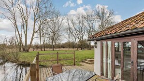 A wooden deck with a table and chairs overlooking a river, with a building featuring large windows in the background. - Pond View Cottage - UK48230 (Tibenham)