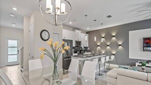 A modern kitchen with white cabinets, a central island, and a dining area with a glass table and white chairs. - Modern Disney Escape - 4BR - Private Pool (Davenport)