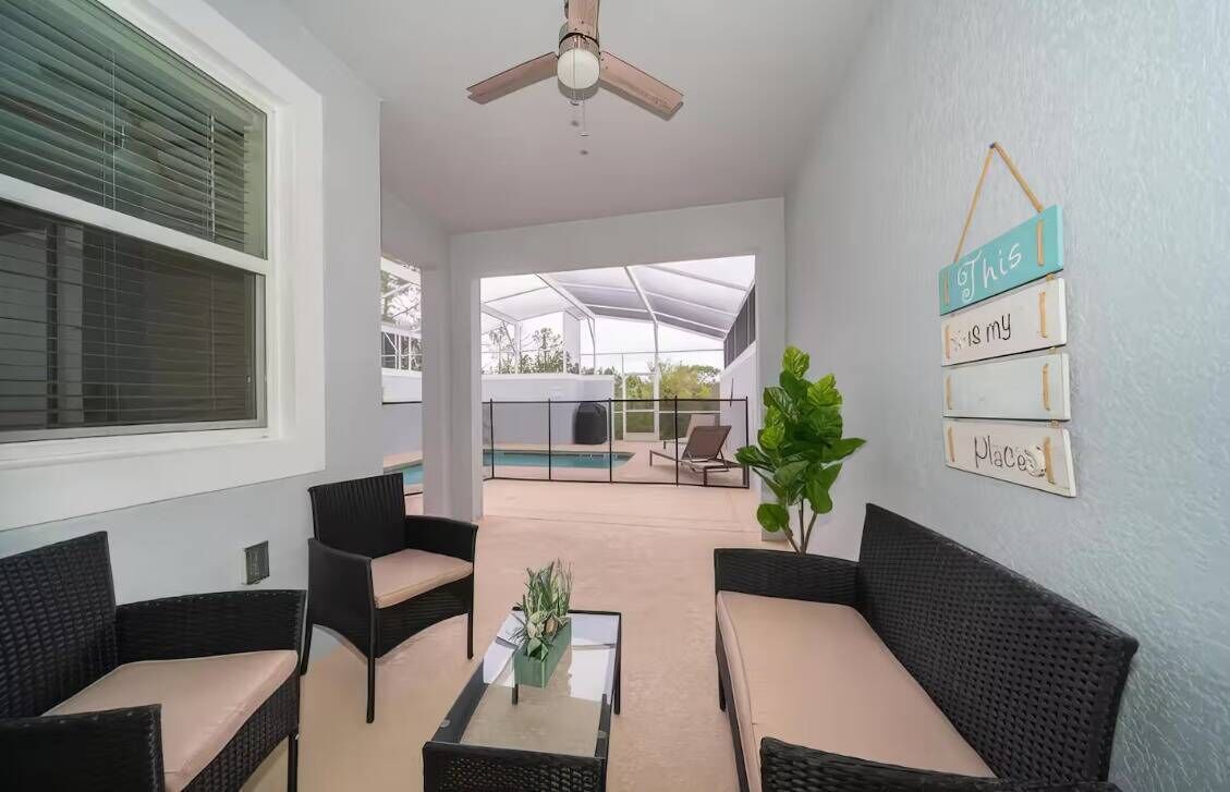 A covered outdoor seating area with wicker furniture and a glass table, a ceiling fan, and a view of a swimming pool.