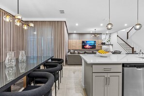 Una cocina moderna con una isla central, un comedor con mesa de vidrio y una sala de estar con sofá y televisor.