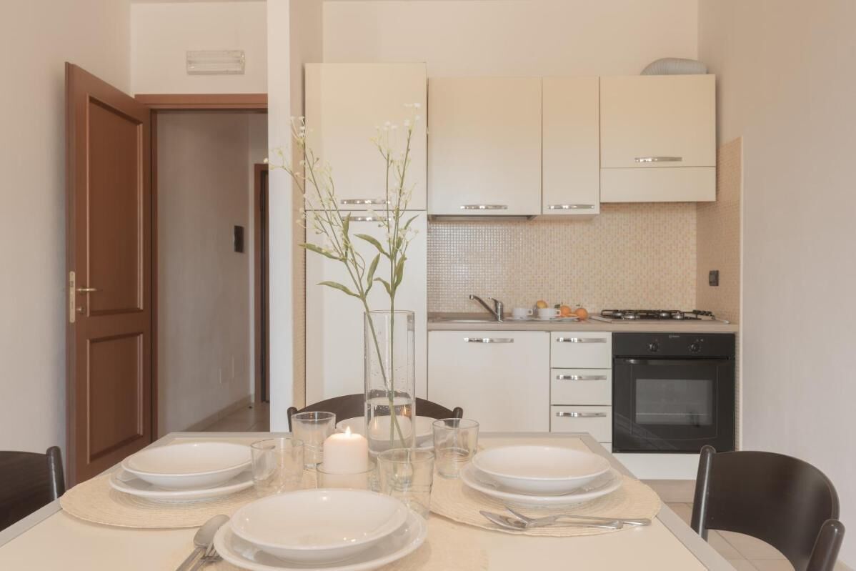 A dining area with a table set for four, a vase with flowers, and a kitchen in the background.