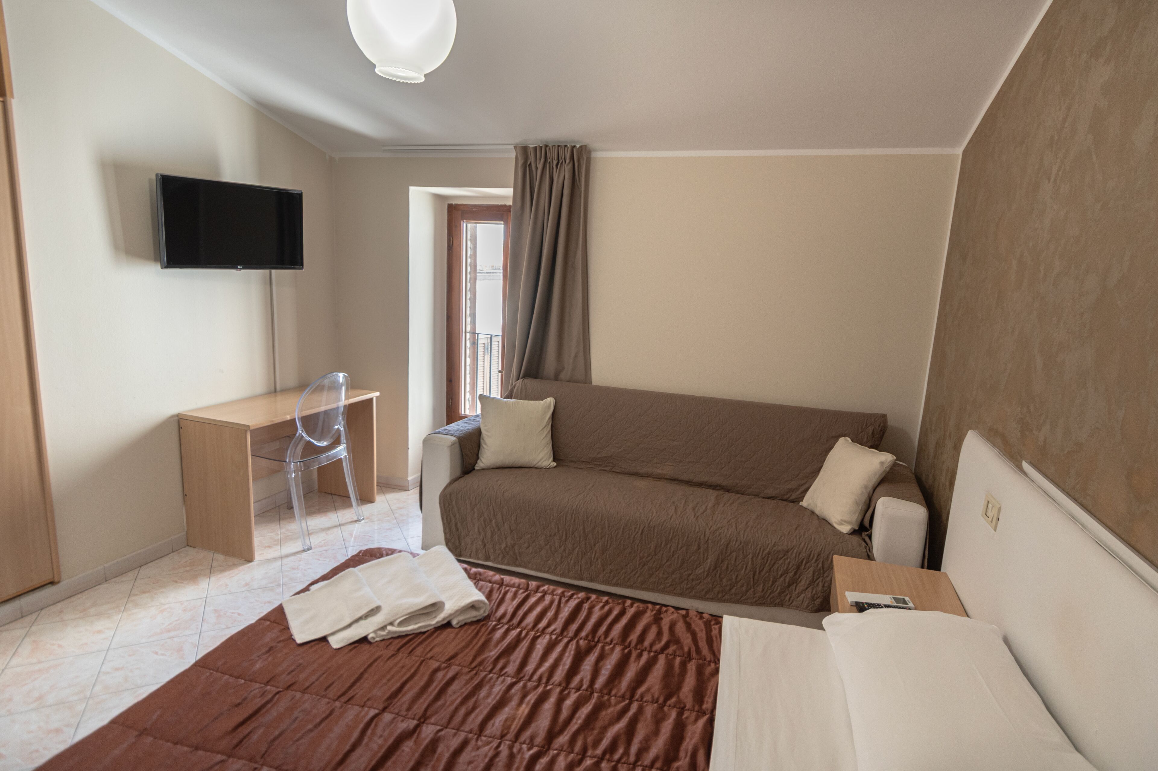 Una habitación de hotel con cama, escritorio, silla, televisión y ventana con cortinas.