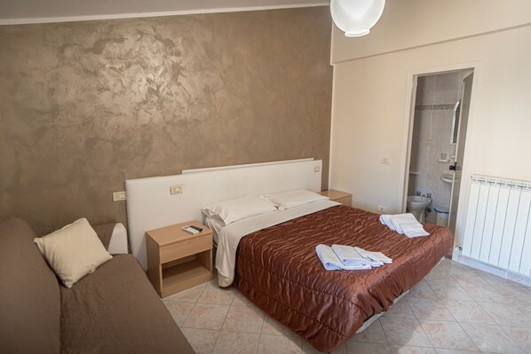 A bedroom with a bed, a nightstand, a bedside table, a bathroom with a sink and shower, and a door leading to another room. - Camere Santa Chiara (Assisi)