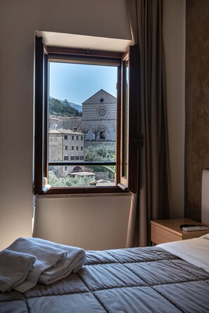 A bedroom with a window, a bed, a nightstand, and a view of a building. - Camere Santa Chiara (Assisi)