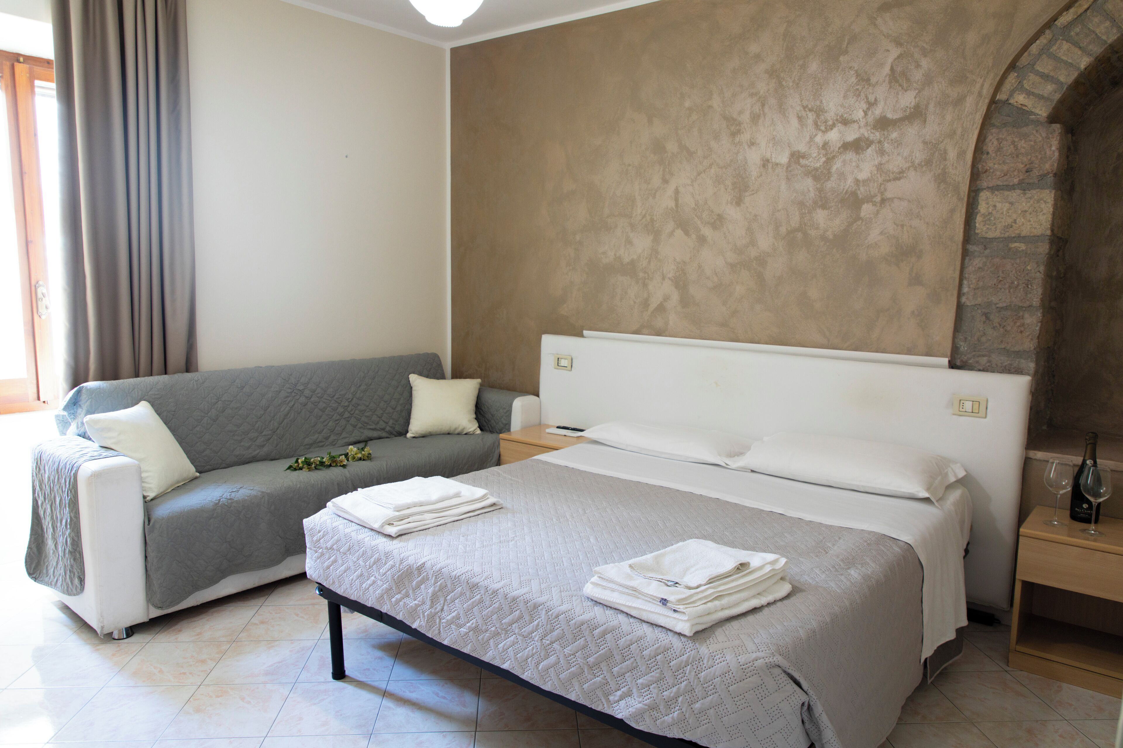 A hotel room with a bed, a sofa, a nightstand, and a stone wall.