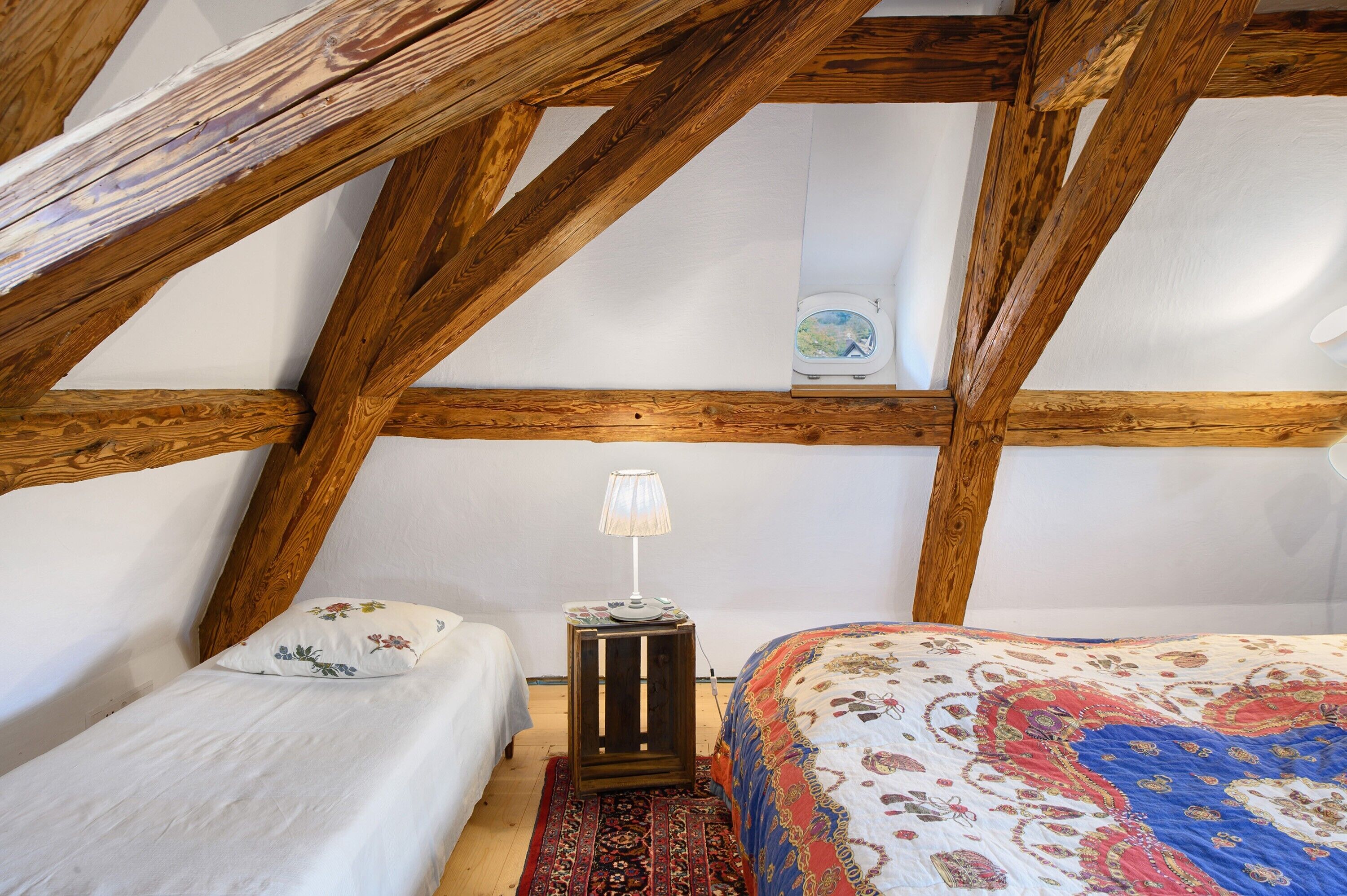 A bedroom with a sloped ceiling, wooden beams, a bed with a patterned bedspread, a bedside table with a lamp, and a small decorative item on the wall.