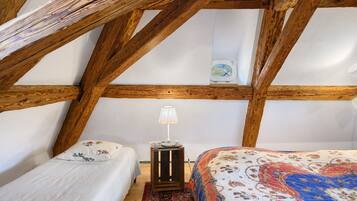 A bedroom with a sloped ceiling, wooden beams, a bed with a patterned bedspread, a bedside table with a lamp, and a small decorative item on the wall.
