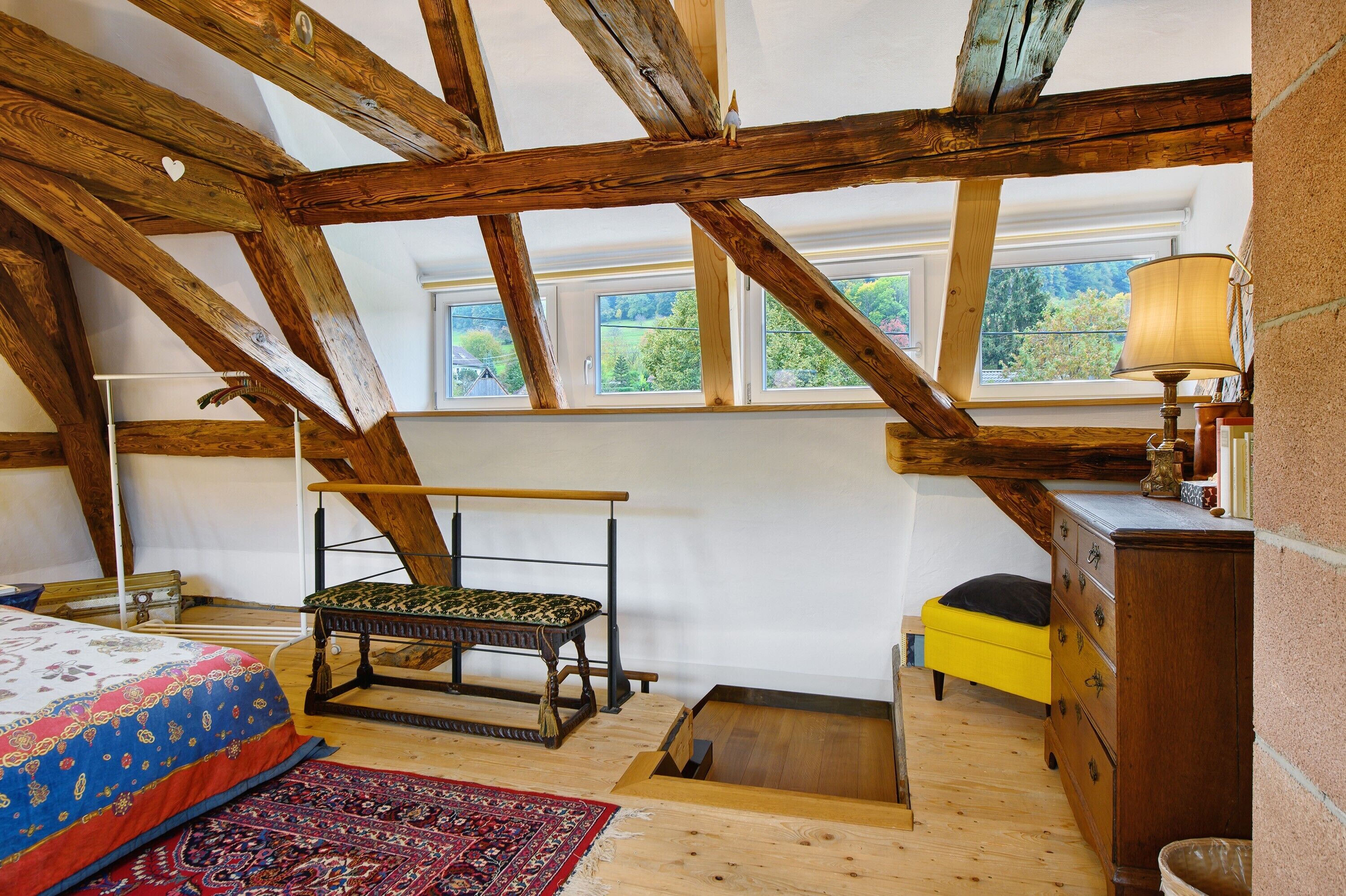 A room with exposed wooden beams, a bed with a patterned bedspread, a bench, a small table with a lamp, and a window with a view of greenery.