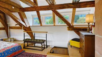 A room with exposed wooden beams, a bed with a patterned bedspread, a bench, a small table with a lamp, and a window with a view of greenery.
