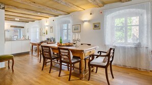 A dining area with a wooden table and chairs, a kitchen in the background, and a window with a view of trees. - Holiday Home 'Zur Alten Dame' with Mountain Views, Private Garden & Wi-Fi (Badenweiler)