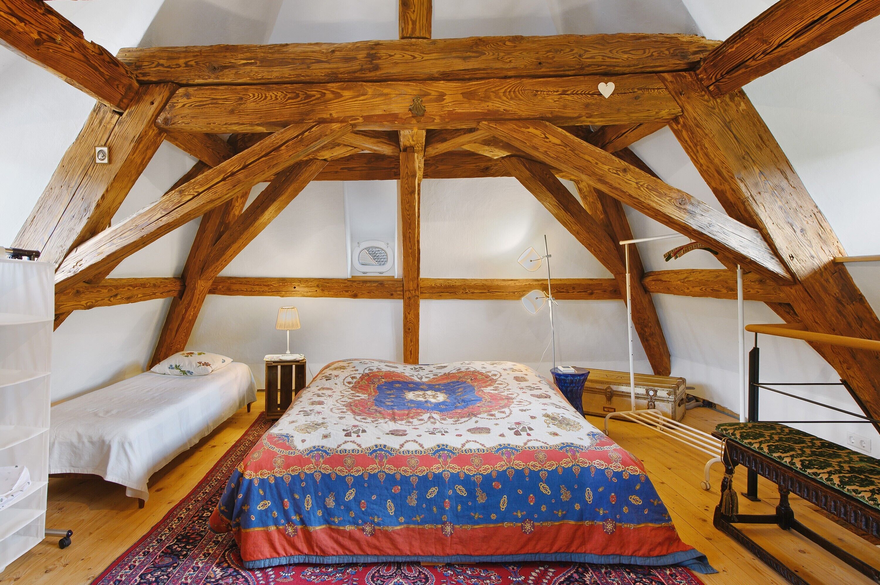 A loft bedroom with a sloped ceiling, wooden beams, a bed with a patterned bedspread, a small bedside table, a lamp, and a suitcase.