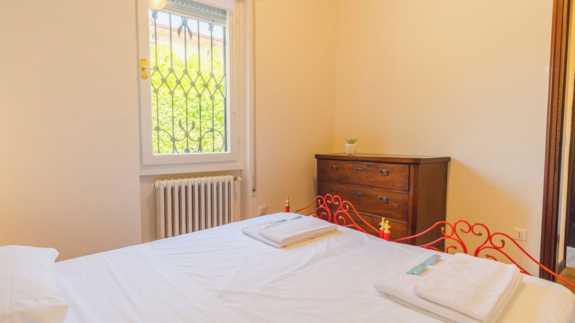 A bedroom with a bed, a radiator, a wooden dresser, and a window with a view of greenery.