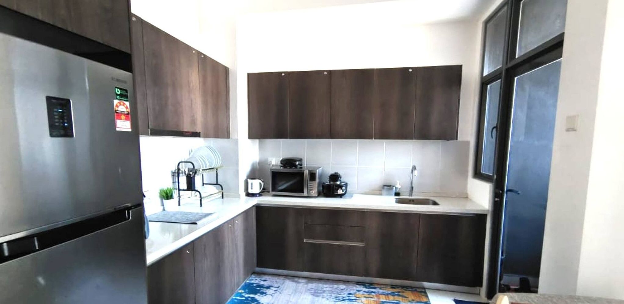 Een kleine keuken met donkerhouten kastjes, een roestvrijstalen koelkast, een magnetron en een spoelbak.