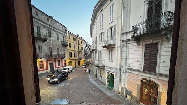 Une rue pavée, bordée de bâtiments des deux côtés, avec un fourgon blanc garé à gauche et une porte verte à droite.